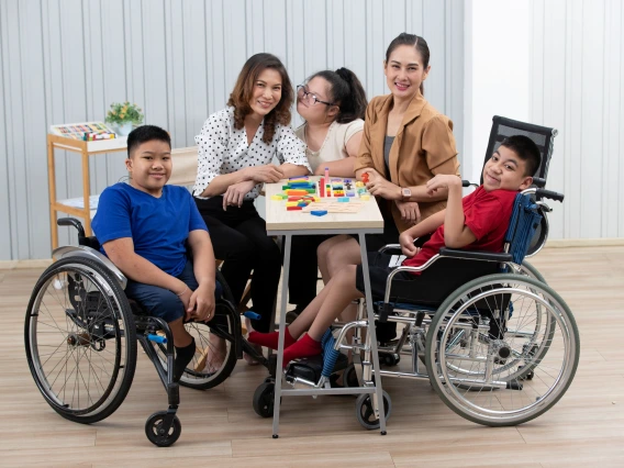 A diverse group of children and educators gather around a table in a classroom, engaging in a hands-on learning activity with colorful blocks. Two children in wheelchairs sit on either side, smiling at the camera, while a young girl with Down syndrome interacts with the teachers. The educators, dressed in business-casual attire, encourage and support the students in an inclusive learning environment.